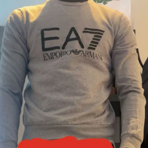 Grå EA7 Emporio Armani sweatshirt - Snygg grå sweatshirt från EA7 Emporio Armani med svart logotyp tryckt på bröstet. Tröjan har rund hals, långa ärmar och är tillverkad i mjuk bomull. Perfekt för dig som gillar stilrena och klassiska märkesplagg med en sportig vibe.