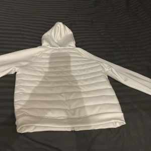 Vit cardigan  - Snygg vit pufferjacka med huva och två praktiska fickor med dragkedja framtill. Jackan har ett quiltat mönster och är tillverkad i ett mjukt syntetmaterial. Perfekt för dig som gillar stilren och clean look. Dragkedja hela vägen framtill för enkel på- och avtagning.