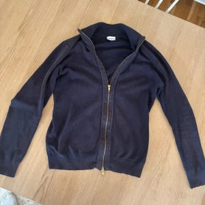Mörkblå kofta från John Henric - Säljer en stilren mörkblå kofta från John Henric i storlek S. Koftan har ett diskret rutmönster, ribbade muddar och krage samt en snygg guldfärgad YKK-dragkedja framtill. Perfekt för lager-på-lager och chill vardagsstil.