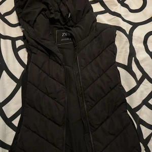 Svart quiltad dunväst från Zara - Svart dunväst från Zara med huva och snyggt quiltat mönster i V-form. Västen har dragkedja framtill och är perfekt för lager-på-lager. Materialet är mjukt och lätt, och designen är stilren med en normal passform.