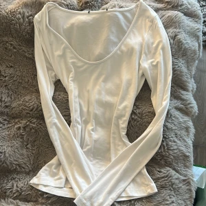 Vit långärmad topp med djup urringning - Säljer en stilren vit långärmad topp med djup rundad urringning. Modellen är slim och sitter snyggt mot kroppen. Perfekt att styla med jeans eller kjol för en clean look. Materialet känns mjukt och skönt mot huden.