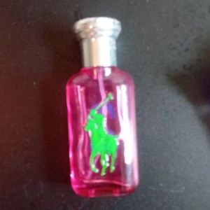 Ralph Lauren rosa parfymflaska - Snygg parfymflaska från Ralph Lauren i transparent rosa glas med en grön polospelare på framsidan. Flaskan har en silvrig kork och en stilren, rektangulär form som känns modern och ungdomlig. Perfekt för dig som gillar färgstarka detaljer och klassisk design. Skulle säga att två tredje delar är andvända. Även haft 2 år så håller absolut länge.