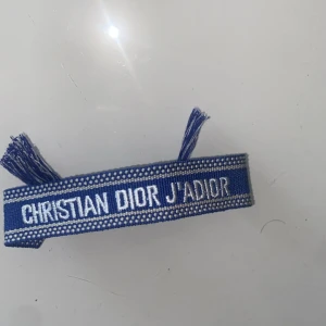 Blått Christian Dior J'Adior armband - Snyggt blått armband från Christian Dior med texten 'J'Adior' broderad i vitt. Armbandet är gjort i vävt tyg med justerbar knytning och fransar i ändarna. Perfekt accessoar för att lyfta din outfit med en lyxig touch.