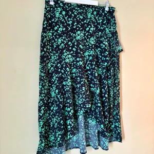 Svart långkjol med grönt blommönster - Flowy svart långkjol med grönt blommigt mönster och asymmetrisk volangkant. Kjolen har omlottmodell med knyt i sidan och är tillverkad i ett lätt och luftigt material som ger snyggt fall.