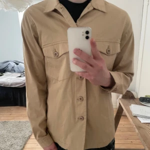 Beige Overshirt från Mango Man - Snygg beige overshirt från Mango Man med långa ärmar och knappar framtill. Skjortan har två bröstfickor med knappar och en klassisk krage. Storlek S. Perfekt för en stilren look nu till våren.