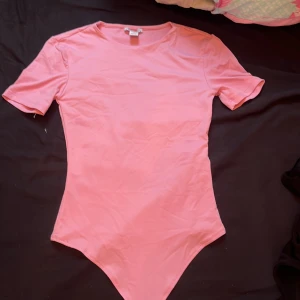 Rosa bodysuit från Amisu XS - Supersöt ljusrosa bodysuit från Amisu i len polyester/satin. Modellen har korta ärmar, rund hals och praktiska tryckknappar i grenen. Formar även midjan.