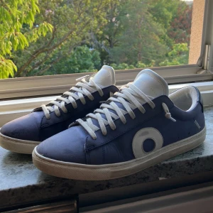  Oliver Cabell low 1 navy  - Säljer ett par stilrena blå sneakers från Oliver Cabell med vit sula och vita snören. Skorna har en rund tå, platt sula och ett stort vitt cirkelmönster på sidan. Ovandelen är i skinn och hälen har en svart detalj med guldfärgad logga. Perfekt för dig som gillar clean design. Ny pris runt 4000 med tull från usa eller Italien 