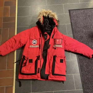 Röd Canada Goose dunjacka med päls - Säljer en röd Canada Goose dunjacka med päls på huvan och flera praktiska fickor. Jackan har reflexdetaljer, svart innerfoder och klassisk patch på bröstet. Perfekt för kalla vinterdagar och har en riktigt schysst look med sin oversized passform.