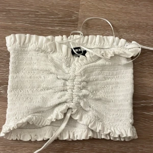 Vit bandeau tubtopp från Bik Bok - Supersöt vit bandeau tubtopp från Bik Bok i storlek S. Toppen har smockad struktur, volangkant upptill och nedtill samt snörning framtill för extra snygg passform. Perfekt för sommarens alla häng och outfits!