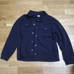 Mörkblå overshirt från Lindex - Snygg mörkblå overshirt från Lindex med klassisk krage och två stora bröstfickor med knappar. Jackan har långa ärmar och stängs med knappar framtill. Perfekt för lager-på-lager och chill stil. Materialet är mjukt och känns skönt mot huden.
