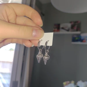 Örhängen med snäckform - Snygga silvriga örhängen med hängande snäckformade berlocker. Örhängena har en rund, klassisk ringdesign och snäckorna är detaljerade och glittrar lätt i ljuset. Perfekt för dig som gillar strandvibbar och coola accessoarer.