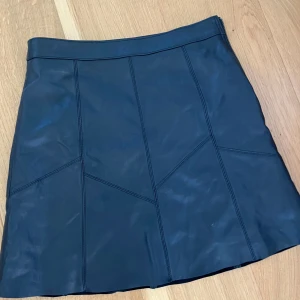 Svart skinnkjol från Odd Molly - Supersnygg svart skinnkjol från Odd Molly med coola sömdetaljer och dragkedja i sidan. Kjolen är kort och har en stilren, lite A-linjeformad passform som ger en edgy vibe. Perfekt att styla med boots eller en stickad tröja för en trendig look. Det är en xs 