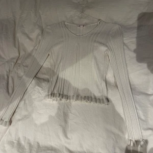 Vit ribbad långärmad tröja - Säljer en vit ribbad långärmad tröja med v-ringad hals och volangdetaljer vid nederkant och ärmslut. Tröjan är stickad i mjukt material och har en slim passform som sitter snyggt på kroppen. Perfekt för dig som gillar stilrena och enkla plagg.