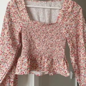 Blommig volangblus med puffärm - Supersöt blus med blommigt mönster i rosa, rött och beige toner. Smockad kropp, volangkant nedtill och långa puffärmar med resår vid ärmslut. Fyrkantig ringning och loose fit över axlarna. 