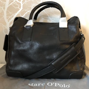 Marc O’Polo svart läderväska  - Stor svart väska i mjukt skinn från Marc O’Polo med silverfärgade metalldetaljer. Väskan har både handtag och justerbar axelrem samt dragkedja upptill. Invändigt finns gott om plats för det du behöver till vardags eller resa. Mått: ca 34 x 37 x 22 cm Skick: Ny, aldrig använd Design: Stilren och tidlös modell i unisex-stil