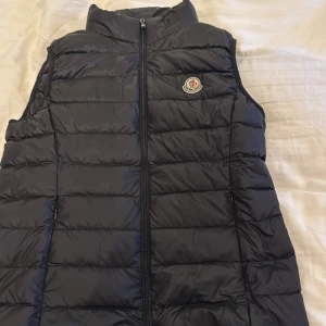 Svart dunväst från Moncler - Snygg svart dunväst från Moncler med quiltad design och klassisk logga på bröstet. Västen har hög krage och dragkedja framtill, perfekt för lager-på-lager. Materialet är glansigt och känns riktigt lyxigt. Passar dig som vill ha en clean och sportig look.