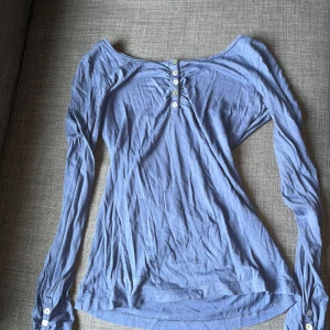 Blå långärmad topp med knappar - Snygg blå långärmad topp med knappar framtill och vid ärmsluten. Toppen har en bred, något offshoulder ringning och är gjord i ett mjukt bomullstyg. Perfekt för dig som gillar en enkel men stilren look.