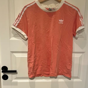 Korallrosa Adidas t-shirt med ränder - Snygg korallrosa t-shirt från Adidas med vita ribbade muddar och klassiska tre ränder längs axlarna. Adidas-logga broderad på bröstet. Skön passform och perfekt för en sportig look. Priset är inte hugget i sten så hör av er om ni har frågor eller liknande😁