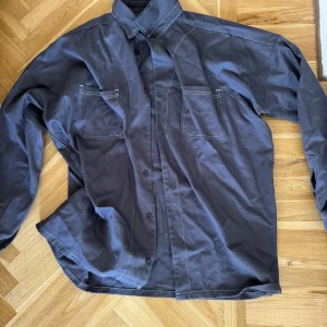 Mörkblå overshirt - Snygg mörkblå skjorta med vita kontrastsömmar och två bröstfickor. Klassisk krage och knäppning framtill. Skjortan har lång ärm och en lite loose passform, perfekt för en avslappnad stil.