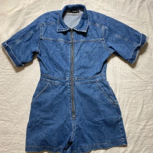 Blå jeansshorts jumpsuit med dragkedja - Snygg blå jeansshorts jumpsuit från Wild Fable med korta ärmar, krage och dragkedja framtill. Klassiska fickor både fram och bak, markerad midja och relaxed fit. Perfekt för en chill och trendig look i sommar.