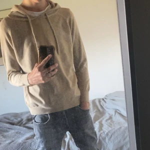 Sand merinoull hoodie  - Beige stickad hoodie med huva och snörning. Den är i färgen sand och har en tajt passform. Perfekt för dig som en enkel och stilren look. Passar till det mesta och är riktigt skön att ha på sig 