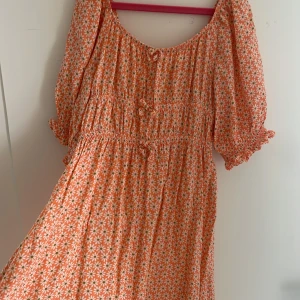 Blommig kortklänning från Zara - Superfin kortklänning från Zara i persikoorange med vita blommor och gröna detaljer. Klänningen har puffiga korta ärmar, smock i midjan och söta rosettdetaljer framtill. Perfekt för sommarens alla tillfällen!