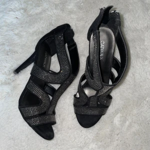 Svarta glittriga klackar från Catwalk - Svarta klackskor från Catwalk med öppen tå och höga klackar. Skorna har snygga remmar täckta av små glittriga stenar och dragkedja bak. Materialet är mockaliknande och designen är riktigt statement. Perfekta för dig som vill sticka ut.