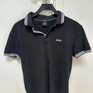 Svart pikétröja från BOSS - Svart pikétröja från BOSS med klassisk krage och vita detaljer på krage och ärmslut. Tröjan har korta ärmar och BOSS-logga broderad på bröstet. Tillverkad i bomull för en skön och stilren känsla.