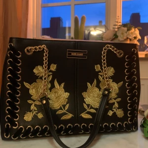 Svart axelväska med guldiga broderier - Snygg svart axelväska från River Island med guldiga kedjehandtag och broderade blommor framtill. Väskan har dekorativa snörningsdetaljer längs kanterna och är tillverkad i skinnimitation. Perfekt för dig som vill sticka ut med en unik accessoar.
