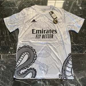 Vit Real Madrid fotbollströja Adidas - Snygg vit Real Madrid fotbollströja från Adidas med svart drakmönster över hela tröjan. Tryck med Emirates Fly Better på bröstet och klubbmärke på vänster sida. Tillverkad i lätt och ventilerande material, perfekt för match eller träning.
