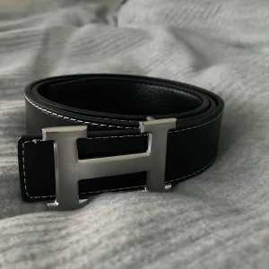 hermesbälte - Säljer ett svart skärp  med ikoniskt H-spänne i silverfärgad metall. Bältet är gjort i äkta skinn och har vita kontrastsömmar längs kanterna. Perfekt accessoar för att lyfta vilken outfit som helst