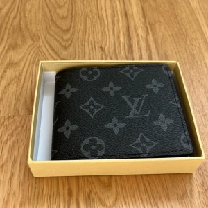 Louis Vuitton svart plånbok i skinn - Snygg svart plånbok från Louis Vuitton med klassiskt monogrammönster i grått. Tillverkad i exklusivt skinn och har flera kortfack samt fönster för ID. Kommer i original ask, perfekt för dig som gillar lyxiga accessoarer.
