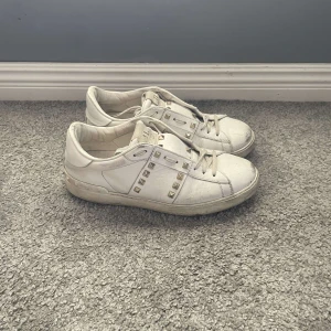 Valentino Rockstud vita sneakers - Snygga vita Valentino Rockstud sneakers i skinn med ikoniska nitar i silver längs sidorna och bak på sulan. Klassisk rund tå, snörning och platt sula. Perfekta för dig som vill ha en lyxig och edgy touch till din outfit.