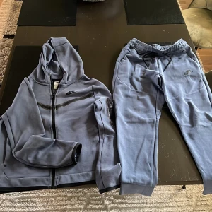 Nike blå mjukisdress med huva - Nike mjukisdress i ljusblått med zip-hoodie och matchande byxor. Hoodien har dragkedja, fickor och svart Nike-logga på bröstet. Byxorna har resår i midjan med snörning och Nike-logga på låret. Perfekt för chill eller träning.