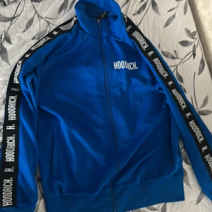 Blå track jacket från Hoodrich - Blå track jacket från Hoodrich med svartvita logoband längs ärmarna och vit tryckt logga på bröstet. Jackan har hel dragkedja, hög krage och två fickor framtill. Perfekt för en sportig och streetinspirerad stil.