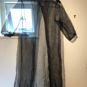 Svart transparent långklänning med paljetter - Snygg svart transparent långklänning i tunn mesh med paljettdekor längst ner. Klänningen har korta ärmar, krage och knäppning framtill. Perfekt för dig som vill sticka ut med en unik och edgy look.
