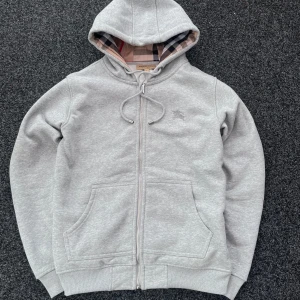 Burberry Zip-Up Hoodie – Grå - 👕 Burberry Zip-Up Hoodie – Grå 📏 Storlek: S 💸 Pris: 900 kr  ✨ Skick & känsla: Extremt skön och snygg hoodie i lyxigt material! I mycket fint skick – inga skador, fläckar eller slitningar. Perfekt till vardags eller för att lyfta en enklare outfit med en touch av designerstil.