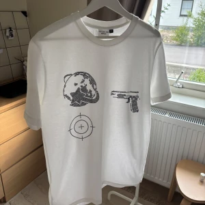 Vit t-shirt Aim For The Moon M - Vit t-shirt från Aim For The Moon i bomull med svart tryck framtill som visar en jordglob, pistol och ett sikte. Klassisk passform med rund hals och korta ärmar.