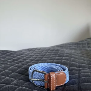 Blått flätat bälte med skinn-detaljer - Snyggt blått bälte med flätad design och bruna skinn-detaljer vid spännet. Spännet är i silverfärgad metall och bältet har en stilren look som passar till jeans eller chinos. Perfekt accessoar för att lyfta din outfit.