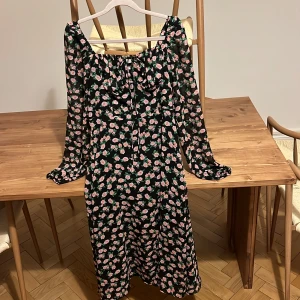 Blommig långklänning från ADOORE - Supersöt långklänning från ADOORE med svart botten och rosa blommor. Klänningen har långa ärmar, snörning vid bysten och en luftig, flowy passform. Perfekt för dig som gillar romantiska vibes och vill sticka ut med ett blommigt mönster.