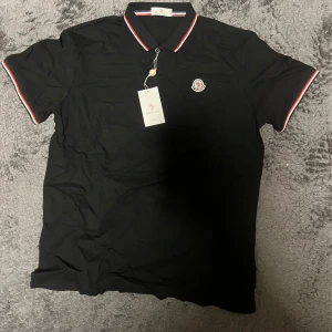 Moncler pike - Svart pikétröja från Moncler med klassisk krage och knäppning. Tröjan har röda och vita ränder på krage och ärmslut samt Moncler-logga på bröstet. Perfekt för dig som gillar stilrena och sportiga plagg med exklusiv känsla. Aldrig använd fick 2 likadana i present så säljer den för lite extra pengar