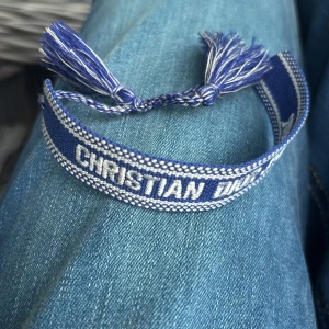 Blått armband från Christian Dior - Snyggt blått tygarmband från Christian Dior med vita detaljer och broderad logga. Armbandet har flätade band och tofsar i ändarna för justerbar passform. Perfekt accessoar för att lyfta din outfit med en lyxig touch.