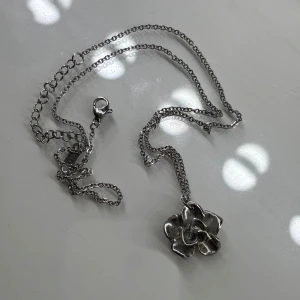 Silverfärgat halsband med blomma K14 - Snyggt halsband i silverfärg med en detaljrik blomformad berlock. Kedjan har ett K14-märke och är justerbar med karbinlås. Perfekt accessoar för att lyfta din outfit med en cool och unik touch.