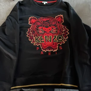 Svart Kenzo sweatshirt med tiger - Svart sweatshirt från Kenzo med ikoniskt broderat tigerhuvud i rött och guld på bröstet. Tröjan har ribbade muddar med gulddetaljer och rund halsringning. Perfekt statement-plagg för dig som vill sticka ut. Storlek S. Kan fraktas men frakten står på er. Priset kan även diskuteras 