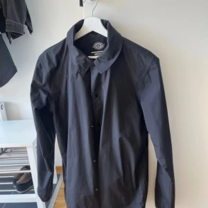 Svart regnjacka från Dickies M - Svart regnjacka från Dickies i storlek medium. Jackan har klassisk krage, knäppning framtill, elastiska muddar och dragsko i midjan