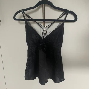 Svart spetsig linnetopp från Censored - Svart linnetopp från Censored med tunna justerbara axelband, spetsdetaljer över bysten och längs sidorna samt liten rosett framtill. Snygg v-ringning och korsade band i ryggen. Toppens material är en mix av satin och spets som ger en lyxig känsla.