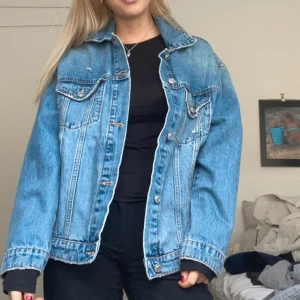 Oversized blå jeansjacka - Säljer en oversized blå jeansjacka med klassisk design, stora bröstfickor med knappar och snygga söm-detaljer. Jackan har lång ärm och knäpps framtill med metallknappar. 