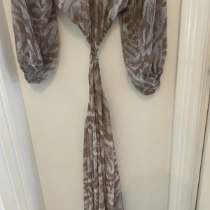 Beige långklänning med mönster - Säljer en beige långklänning med vitt abstrakt mönster. Klänningen har långa, transparenta ärmar och en snygg omlottknytning i midjan. Materialet är lätt och flowy, perfekt för varma dagar. Djup v-ringning och lös passform ger en chill vibe.