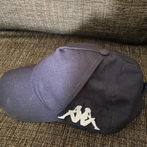 Mörkblå keps från Kappa - Snygg mörkblå keps från Kappa med det klassiska vita Kappa-loggan broderad på sidan. Kepsen är tillverkad i 100% bomull och har en klassisk böjd skärm. Perfekt accessoar för en sportig och avslappnad stil.