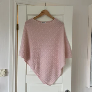 Ljusrosa kashmirponcho från Davida - Supermjuk ljusrosa ponchotröja från Davida i 100% kashmir. Tröjan har ett snyggt kabelstickat mönster och en lös passform med rund hals. 